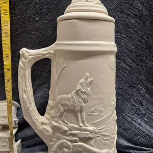May include: White ceramic beer stein with a wolf howling at the moon design. The stein has a handle shaped like a tree branch and a lid with a wolf on top.