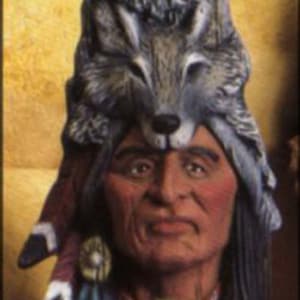 May include: A ceramic figurine of a Native American man wearing a headdress with two wolf heads. The headdress is made of brown, gray, and white ceramic. The man has a brown face and black hair. The figurine is sitting on a wooden base.