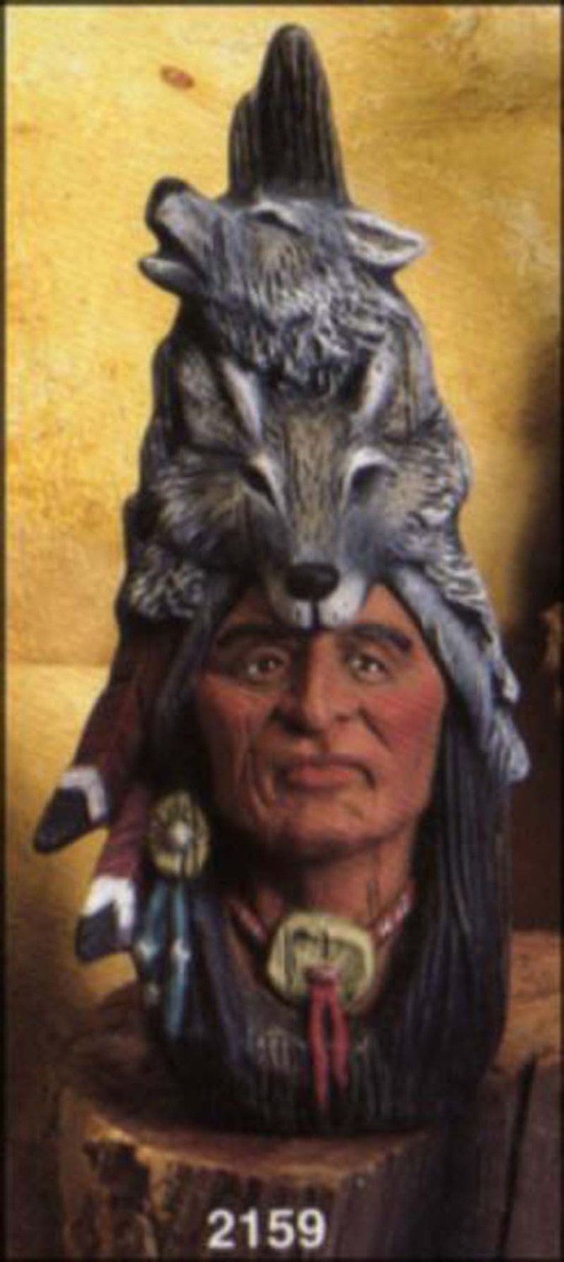 May include: A ceramic figurine of a Native American man wearing a headdress with two wolf heads. The headdress is made of brown, gray, and white ceramic. The man has a brown face and black hair. The figurine is sitting on a wooden base.