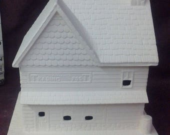 May include: White ceramic unpainted miniature house with a shingled roof and a sign that reads "Trading Post".