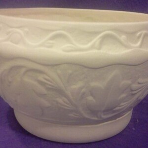 May include: A white ceramic planter with a wide, rounded shape. The planter features a decorative embossed design of leaves and a wavy pattern around the top. The base is a smaller, rounded pedestal.
