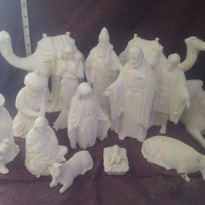 May include: A collection of unpainted ceramic figurines depicting a nativity scene. The set includes figures of the three wise men, Mary, Joseph, baby Jesus in a manger, camels, a cow, and other figures. The figurines are all white and unpainted.