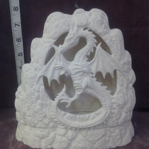 May include: Unpainted ceramic dragon figurine, approximately 7 inches tall. The dragon is sculpted with detailed wings and scales, set within a rocky, cave-like structure. The piece is designed to hold a light source.