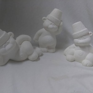 May include: Three unpainted ceramic bear figurines. Each bear wears a hat with a textured design. One bear is lying down, another is standing, and the third is sitting. The bears have a smooth, matte finish and are ready for painting.