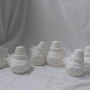 May include: Six white ceramic snowman figurines with painted-on facial features. Each snowman has a hat and a scarf. The snowmen are arranged in a row on a white surface.