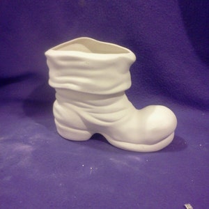 May include: White ceramic boot shaped planter with a folded top. The boot is sitting on a purple surface.