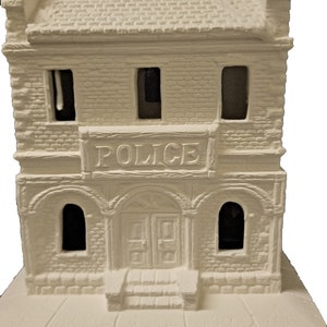 May include: White ceramic model of a police station with a sign that reads "POLICE" above the entrance. The building has two windows on the top floor and two arched windows on the bottom floor. There are steps leading up to the entrance.