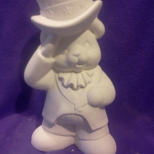 May include: A white ceramic figurine of a rabbit dressed in patriotic attire. The rabbit wears a top hat with stars, a vest, and a ruffled collar. The figurine is unpainted and ready for customization.