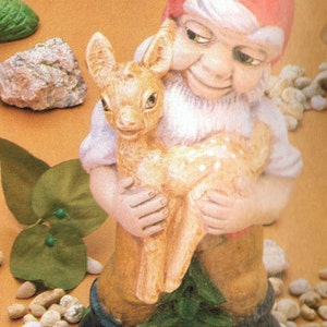 May include: A ceramic gnome figurine holding a fawn. The gnome is wearing a red hat and has a white beard. The fawn is brown with white spots. The figurine is 13 inches tall. DH258 - Elf Holding Fawn 13" T