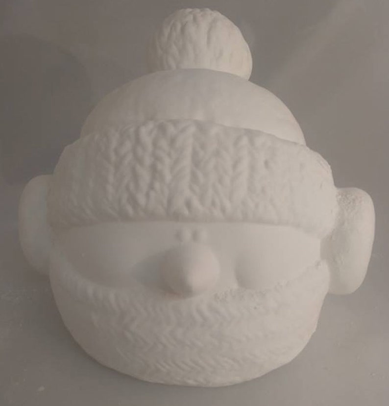Op de afbeelding: Witte keramische figuur van een persoon met een gebreide muts en een sjaal. De figuur heeft een rond gezicht met een kleine neus en een brede glimlach.