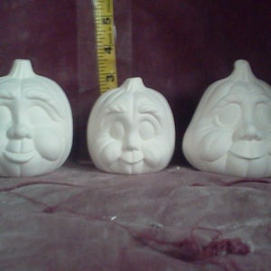 May include: Three white ceramic pumpkins with painted faces. Each pumpkin has a different facial expression. The pumpkins are all approximately the same size.