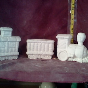 May include: A set of three white ceramic train cars, ready to be painted. The cars are a locomotive, a boxcar, and a caboose.