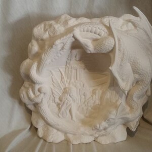 May include: White ceramic wall plaque with a raised design of two dragons surrounding a castle. The dragons are in a curled position with their wings spread out. The castle is in the center of the plaque and has a tower and a gate.