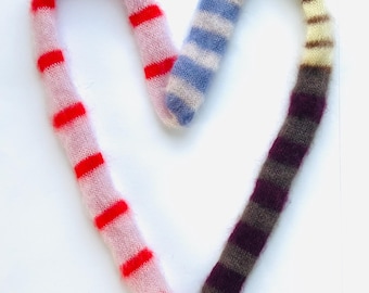 May include: A heart-shaped, knitted item with a fluffy texture. The left side is light pink with red stripes, transitioning to blue and white stripes, and ending with purple and brown stripes. The item is on a white background.