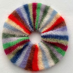 May include: A colorful, handmade scrunchie with a striped pattern. The scrunchie features a variety of colors, including red, green, blue, purple, and yellow. The fabric appears soft and fluffy, perfect for accessorizing a hairstyle.