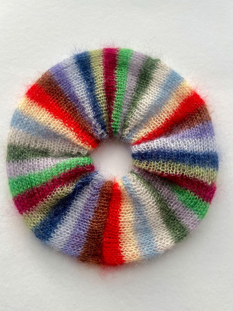 May include: A colorful, handmade scrunchie with a striped pattern. The scrunchie features a variety of colors, including red, green, blue, purple, and yellow. The fabric appears soft and fluffy, perfect for accessorizing a hairstyle.