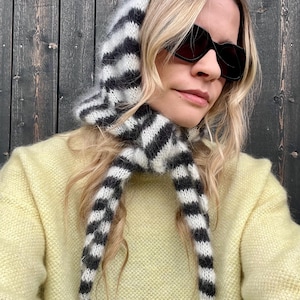 Könnte beinhalten: Eine schwarz-weiß gestreifte Strickhaube mit langen Bindebändern, getragen über einem gelben Pullover. Die Kapuze ist aus einem weichen, flauschigen Material gefertigt. Die Person trägt eine schwarze Sonnenbrille.