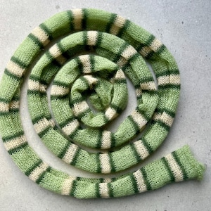 May include: A knitted scarf in a spiral shape, featuring alternating bands of light green and off-white. The scarf is made of a soft, textured yarn, and the design is visible from all angles. The scarf is a unique accessory.