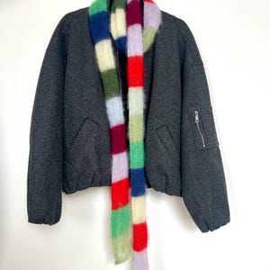 Könnte beinhalten: Eine dunkelgraue Bomberjacke mit einem bunten, gestreiften Schal. Der Schal hat vertikale Streifen in Grün, Blau, Rot, Lila und Weiß. Die Jacke hat einen Reißverschluss und zwei Vordertaschen.