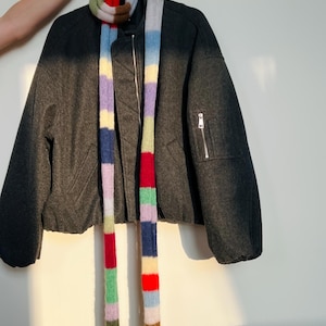 May include: A dark gray bomber jacket with a colorful striped scarf. The scarf has vertical stripes of red, blue, green, purple, and yellow. The jacket has a zipper closure and a small pocket on the sleeve.