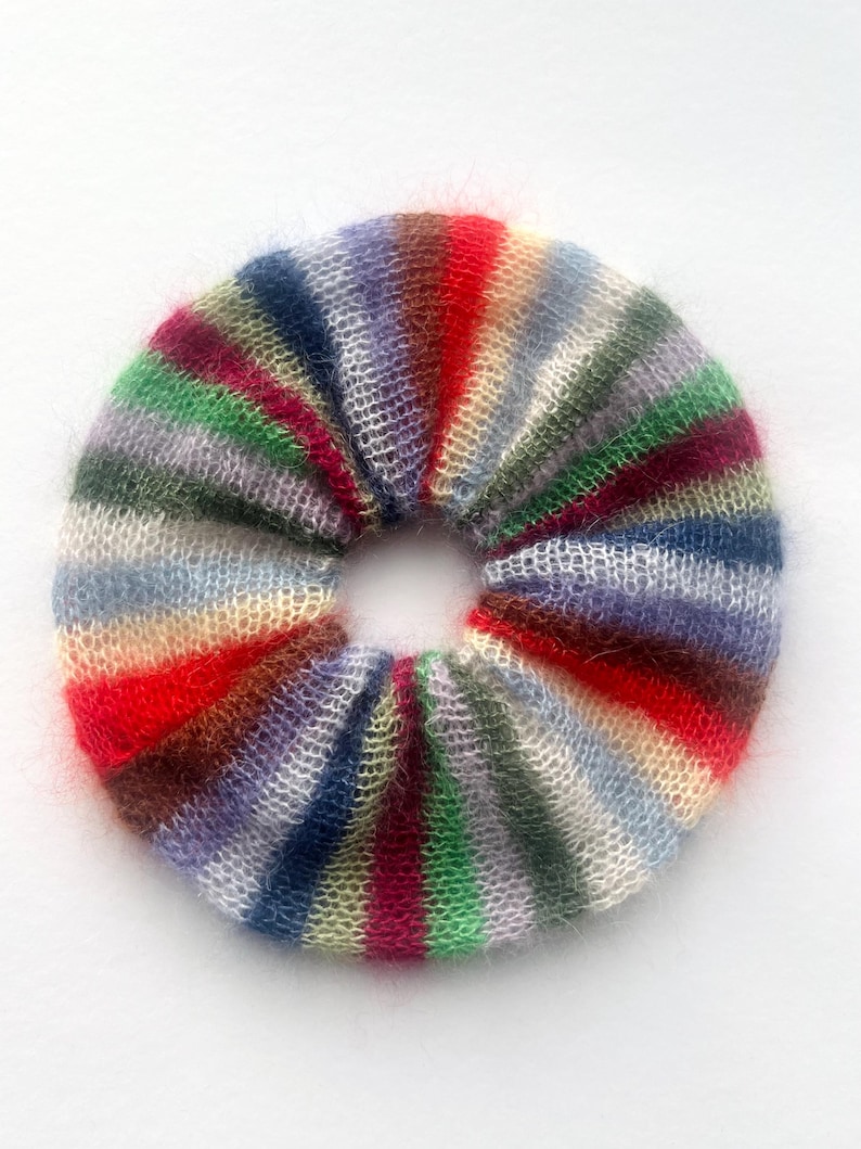 May include: A colorful, striped scrunchie in a circular shape. The scrunchie features a variety of colors, including red, green, blue, and purple. The material appears to be soft and fuzzy, with a knitted or woven texture. This hair accessory is perfect for adding a pop of color to any hairstyle.