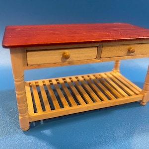 May include: A miniature wooden kitchen island with a dark red tabletop and two small drawers. The island has a slatted shelf and is supported by light-colored, turned legs. The drawer pulls are small, round, and gold-colored.