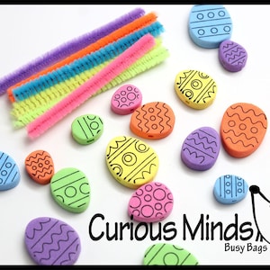 以下が含まれることがあります： 黒と白のパターンが描かれたカラフルなフォーム製のイースターエッグビーズのセット。ビーズはパイプクリーナーに通されています。画像には「Curious Minds Busy Bags」というテキストも含まれています。
