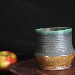 May include: A handmade ceramic mug with a dark gray exterior, a textured, speckled orange base, and a teal green rim. A red and yellow apple sits to the left of the mug on a wooden surface, against a dark background. The mug is approximately 4 inches tall.