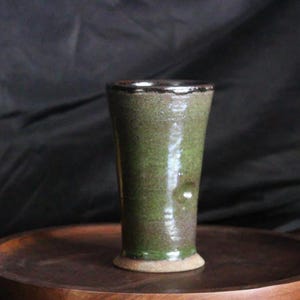 May include: A handmade, green and brown ceramic tumbler with a textured surface. The tumbler has a flared shape and a dark rim. It sits on a wooden tray, with a red and yellow apple in the background.