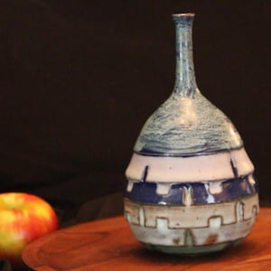 May include: A ceramic vase with a long, slender neck and a rounded body. The upper portion features a mottled blue and white glaze, while the lower section displays a geometric pattern of white, dark blue, and tan bands with vertical indentations.