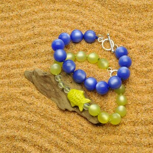 May include: Two beaded necklaces, one blue and one yellow, lay on a sandy surface. The blue necklace has a silver clasp and is made of round beads. The yellow necklace is made of smaller, round beads. A yellow fish charm hangs from the yellow necklace.