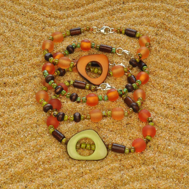 May include: Three layered necklaces with brown wooden beads, orange glass beads, and green glass beads. The necklaces feature two large wooden pendants with a green and orange color scheme.
