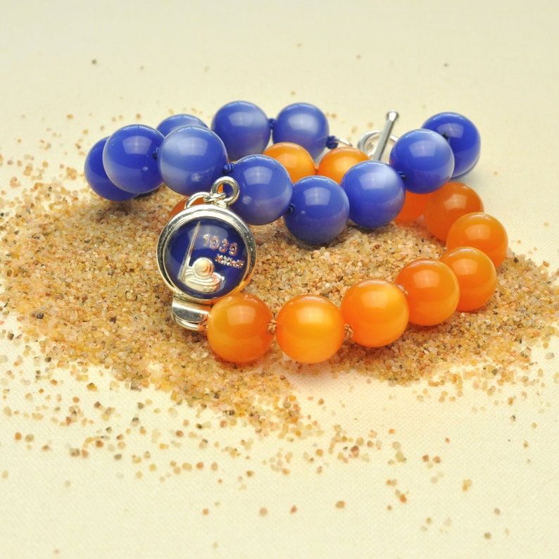 May include: Two bracelets, one with blue beads and one with orange beads, are lying on a sandy surface. A silver charm with the text "1939" is attached to the blue beaded bracelet.