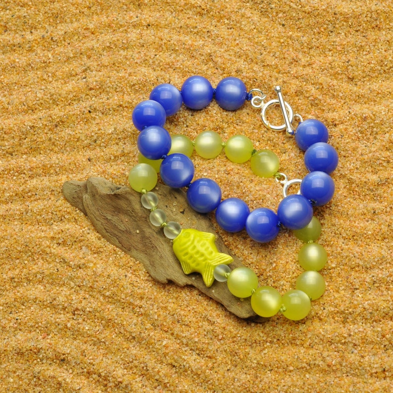 May include: Two beaded necklaces, one blue and one yellow, lay on a sandy surface. The blue necklace has a silver clasp and is made of round beads. The yellow necklace is made of smaller, round beads. A yellow fish charm hangs from the yellow necklace.