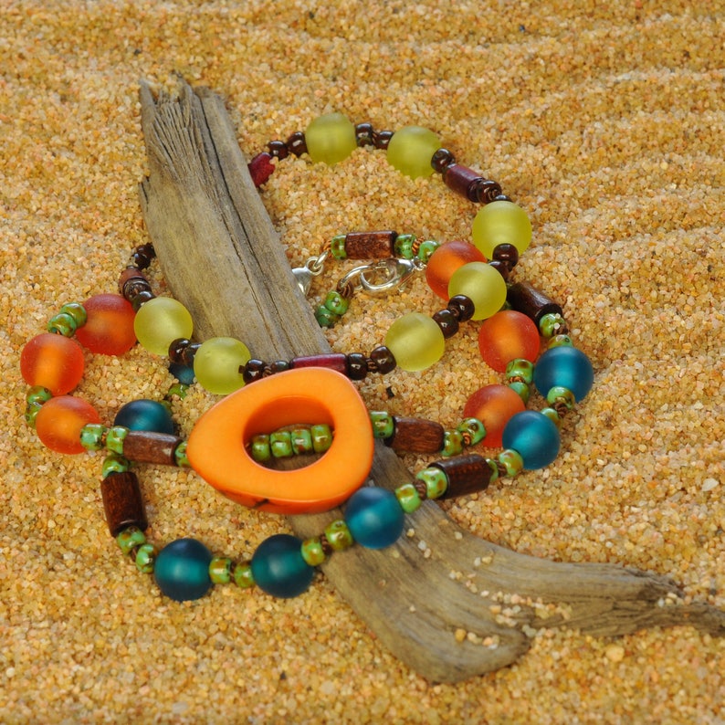 May include: A colorful necklace with orange, green, blue, and brown beads on a driftwood background. The necklace has a large orange pendant with a hole in the center.