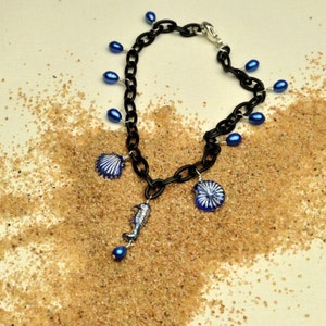 May include: A black chain necklace with blue glass beads and silver charms shaped like a seashell, a seahorse, and a spiral shell. The necklace is lying on a pile of sand.