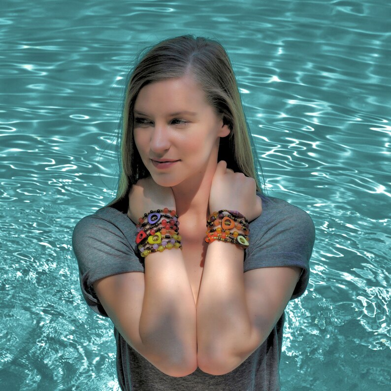 May include: A woman wearing a gray shirt with colorful bracelets on both wrists. The bracelets are made of beads and have a variety of shapes and colors.