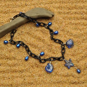 May include: A black chain necklace with blue glass beads and charms. The charms are shaped like seashells and a starfish. The necklace is lying on a sandy surface.