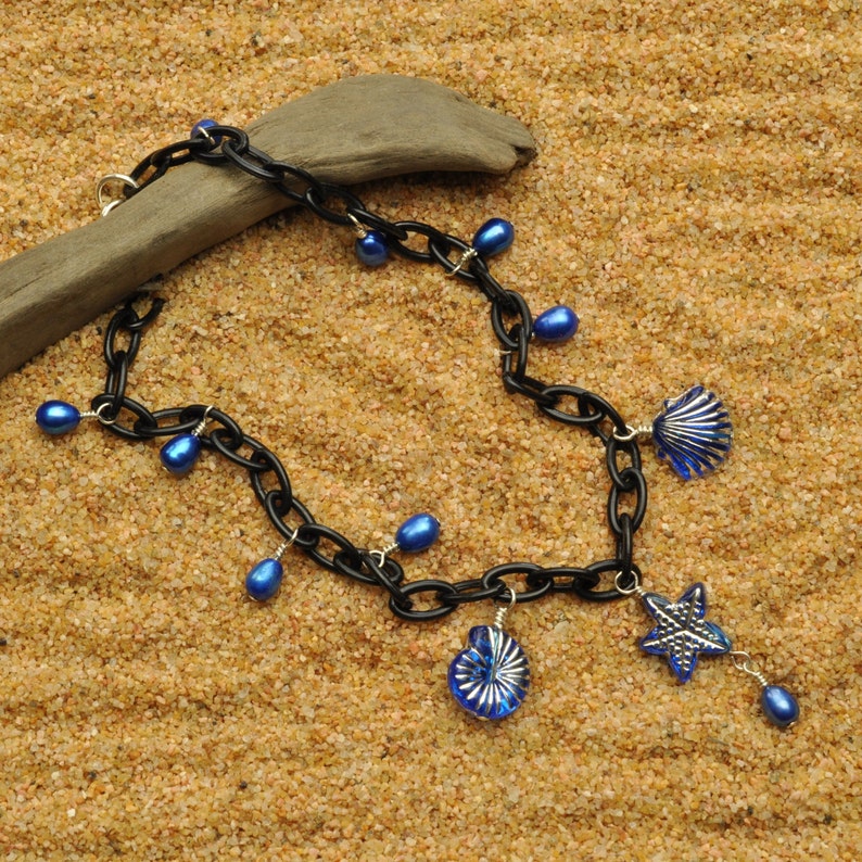 May include: A black chain necklace with blue glass beads and charms. The charms are shaped like seashells and a starfish. The necklace is lying on a sandy surface.