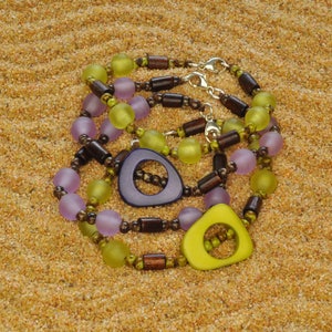 May include: Three layered necklaces with wooden beads, purple and green glass beads, and two large wooden pendants. The pendants are shaped like a circle with a hole in the middle. One pendant is purple and the other is green.