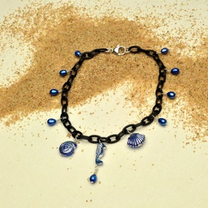 May include: A black chain necklace with blue glass beads and silver charms shaped like seashells and a seahorse. The necklace is on a white background with sand.