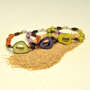 May include: Three colorful bracelets with wooden beads and geometric shaped charms. The bracelets are arranged on a pile of sand.