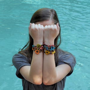 May include: A woman with long brown hair is wearing a stack of colorful beaded bracelets on both wrists. The bracelets are made of various colors, including blue, green, yellow, and orange. The bracelets are made of small, round beads that are strung together on a thin cord.