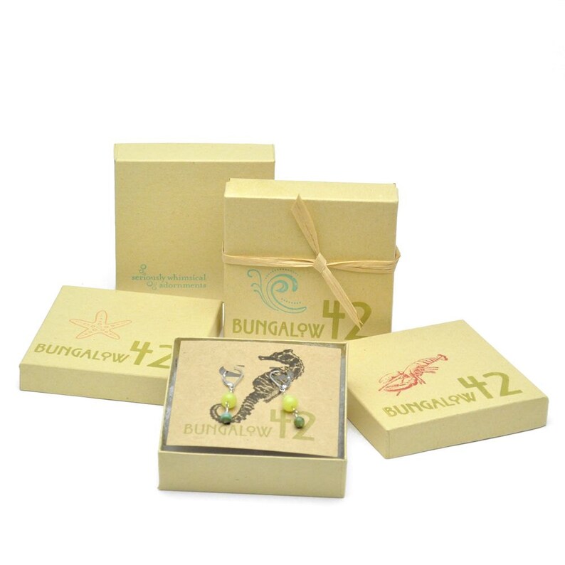 May include: A set of green and yellow gemstone earrings in the shape of a seahorse, packaged in a small brown box with the text "Bungalow 42" printed on it.