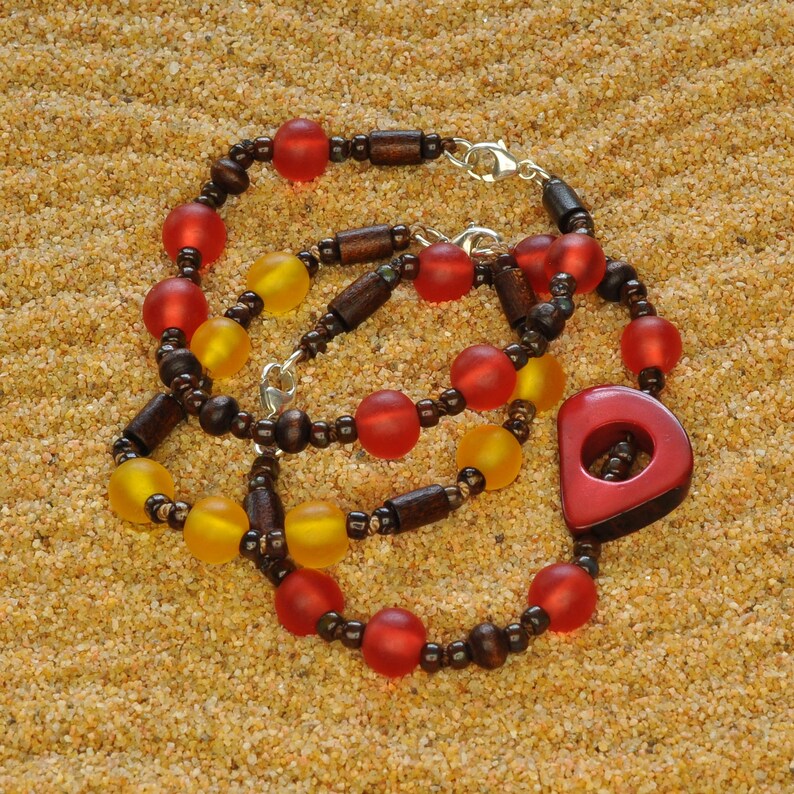 May include: A layered necklace with wooden beads, red and yellow glass beads, and a large red wooden pendant. The necklace is on a sandy surface.