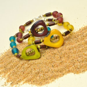 May include: Three colorful bracelets with wooden beads and geometric shaped pendants in shades of brown, yellow, and green. The bracelets are on a bed of sand.