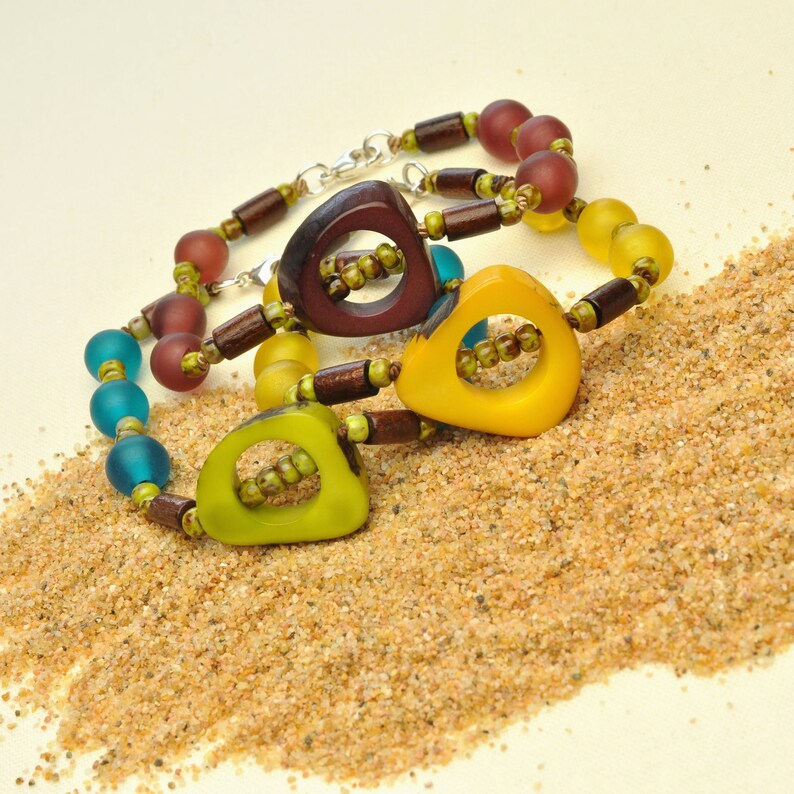 May include: Three colorful bracelets with wooden beads and geometric shaped pendants in shades of brown, yellow, and green. The bracelets are on a bed of sand.