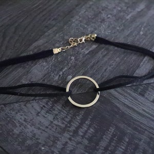May include: A black choker necklace with a gold-colored circular pendant. The necklace features a thin, black suede-like band and a gold-colored chain clasp for adjustable sizing. The pendant is a simple, elegant circle.