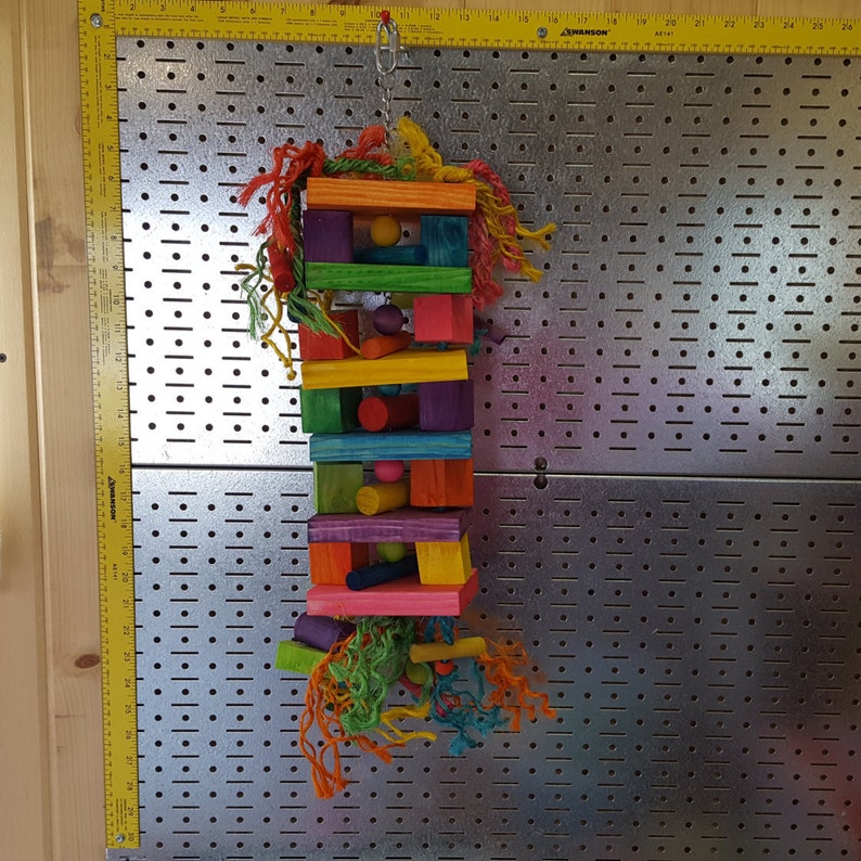 May include: A colorful wooden bird toy with multiple layers of brightly colored wood blocks, beads, and ropes. The toy is hanging from a metal hook.