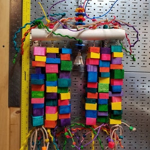 May include: A colorful wooden bird toy with multiple hanging blocks, beads, and strings. The toy is suspended from a white PVC pipe and has a bell hanging from the center.